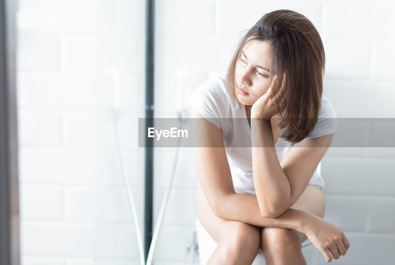 Thoughtful beautiful woman sitting on toilet bowl at home