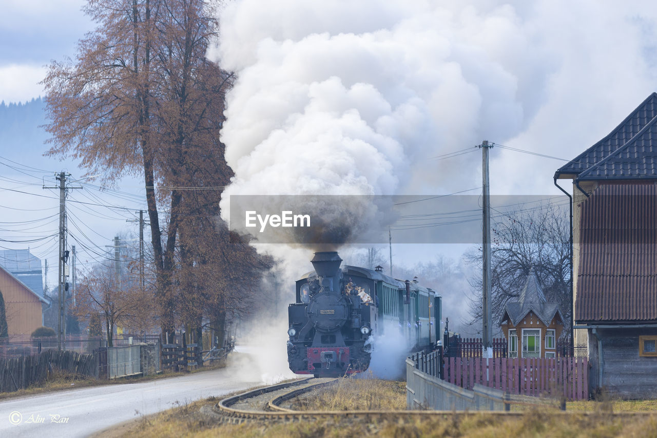 Front view of a steam train running on a railtrack bext to a road and houses