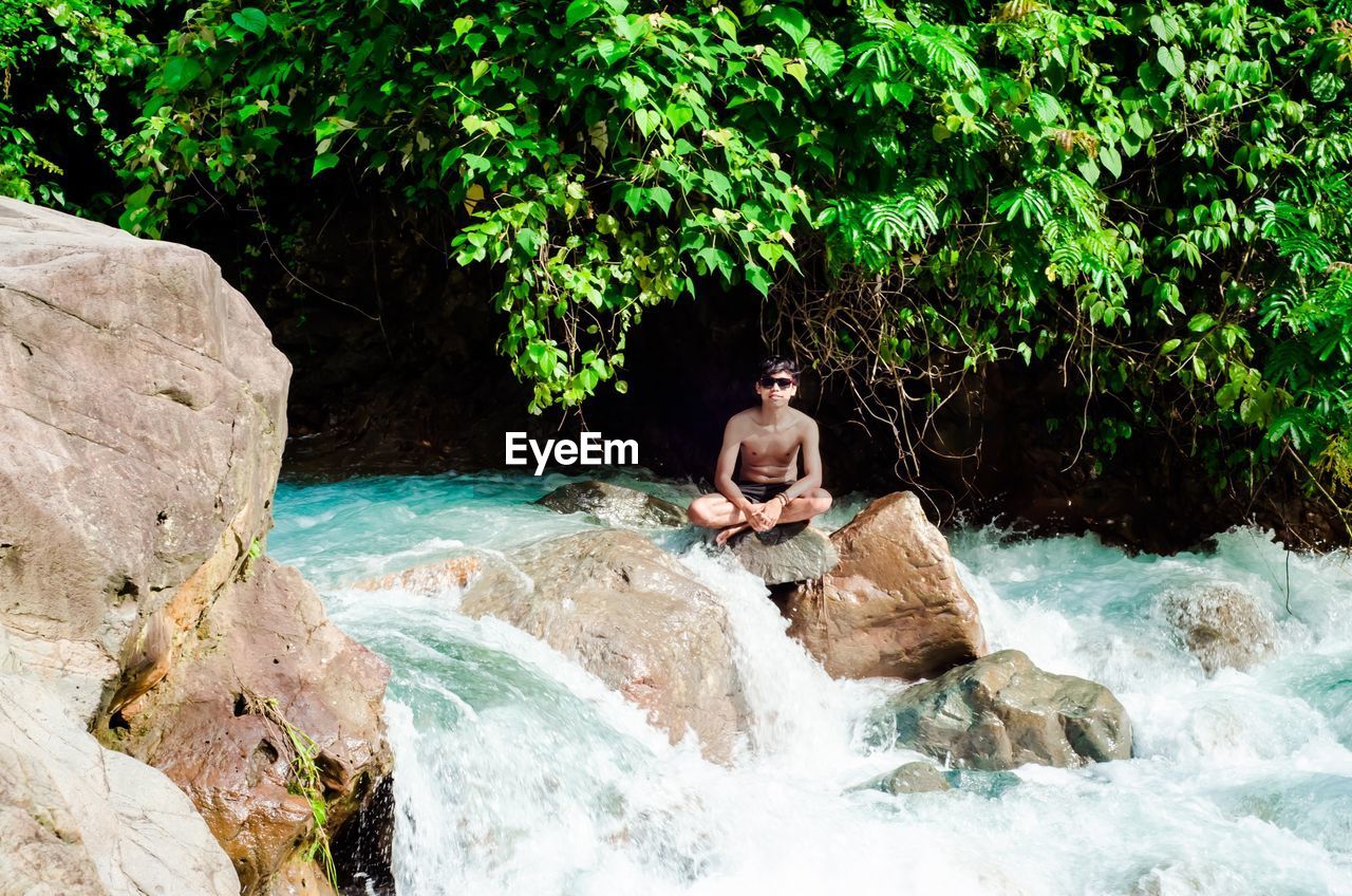 Full length of shirtless man sitting on rock by river in forest