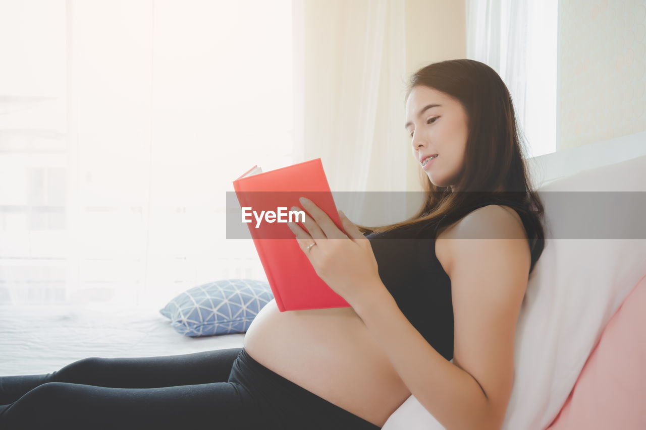 Side view of pregnant woman reading book while sitting on bed at home