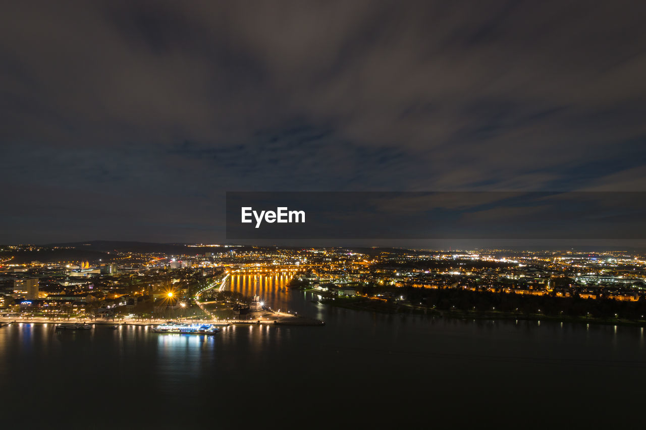 ILLUMINATED CITY BY RIVER AGAINST SKY AT NIGHT