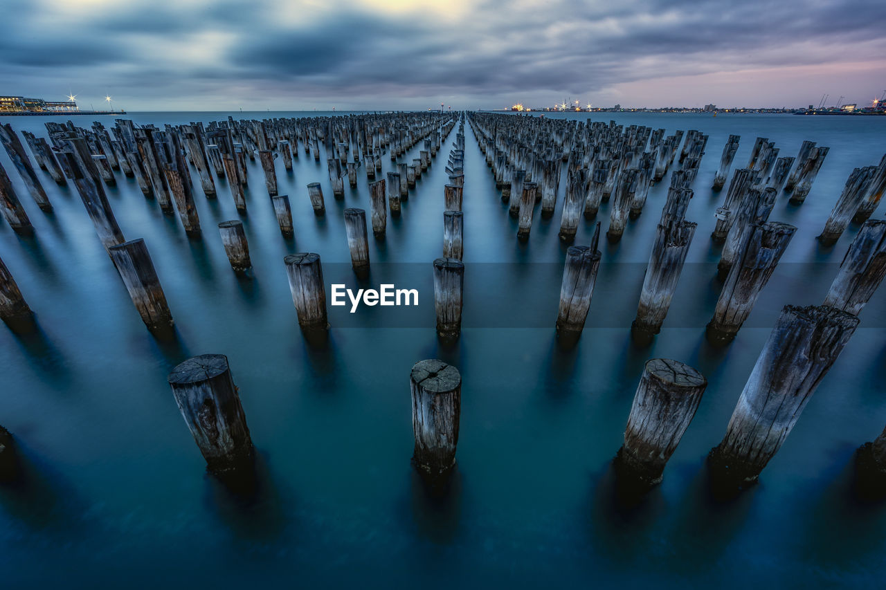 Wooden posts in sea against cloudy sky during sunset