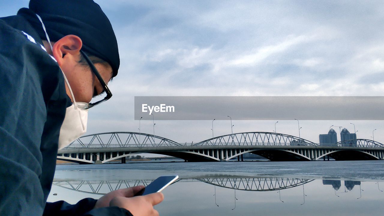 Man wearing mask using mobile phone against bridge over river