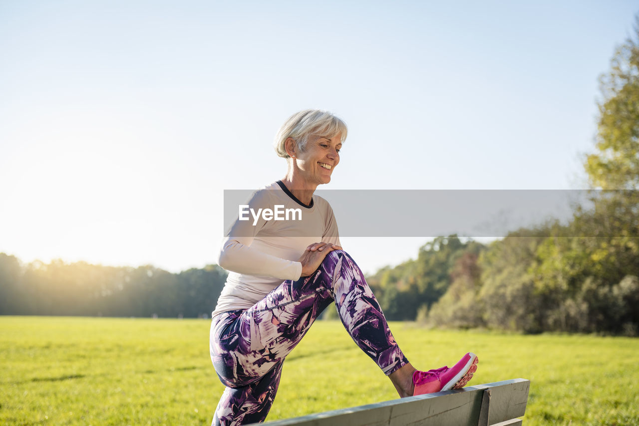 Smiling senior woman stretching on a bench in rural landscape