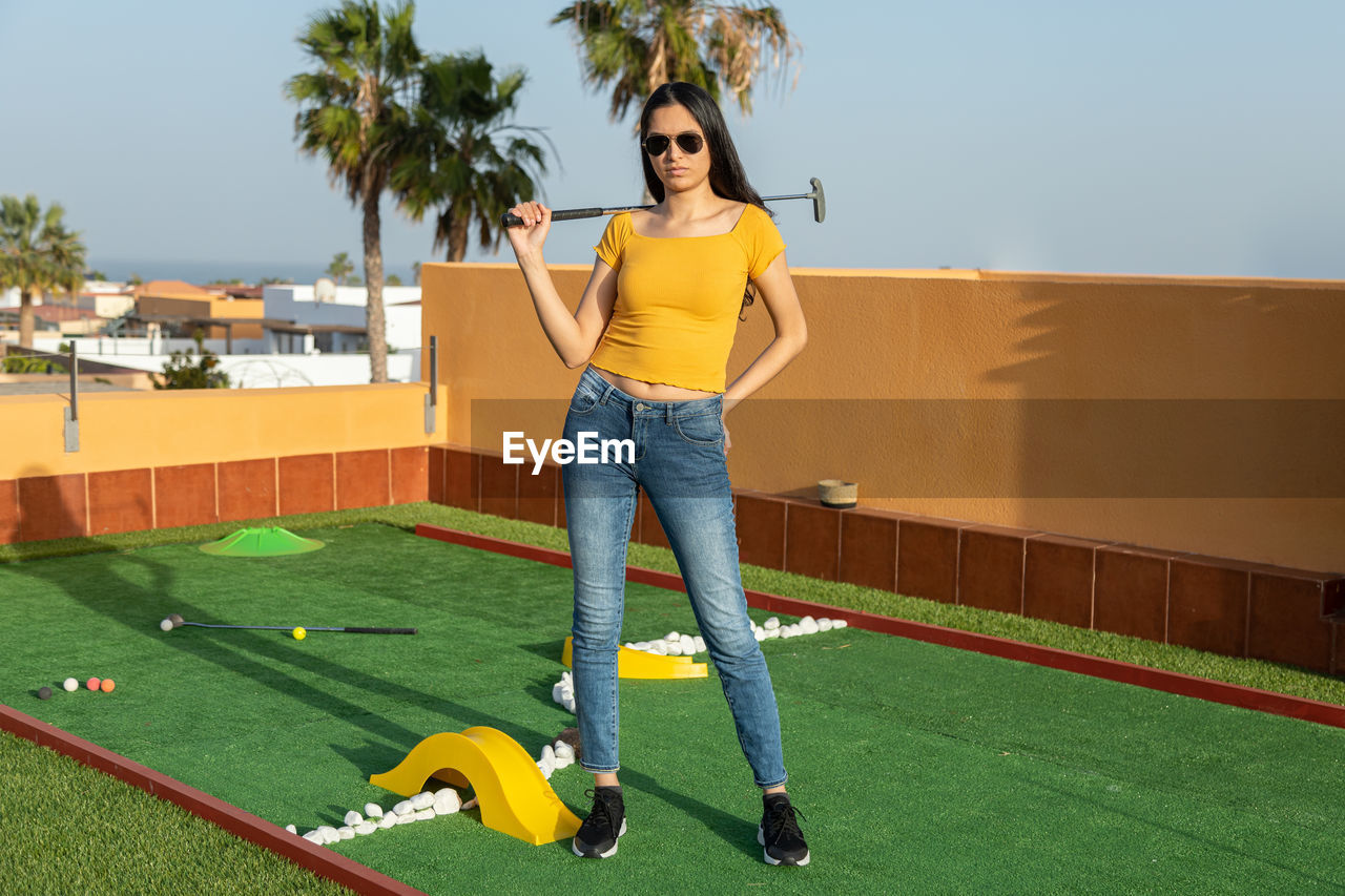 one person, full length, adult, sports, grass, leisure activity, young adult, palm tree, casual clothing, plant, women, miniature golf, lifestyles, nature, emotion, standing, smiling, happiness, person, yellow, portrait, day, tropical climate, playground, front view, tree, architecture, clothing, fun, outdoors, hairstyle, looking at camera, brown hair, sport venue, green, activity, copy space, enjoyment