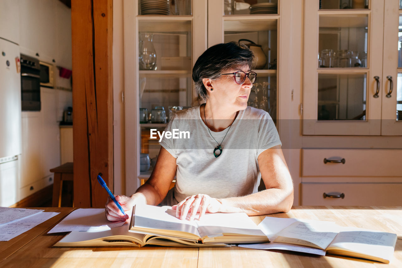 Focused middle aged female in glasses with pen in hand sitting at table with heap of books and papers and studying professional materials at home