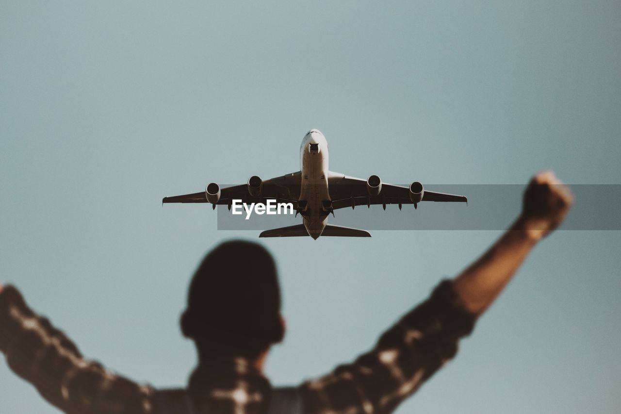 Rear view of man with arms outstretched against airplane flying in sky