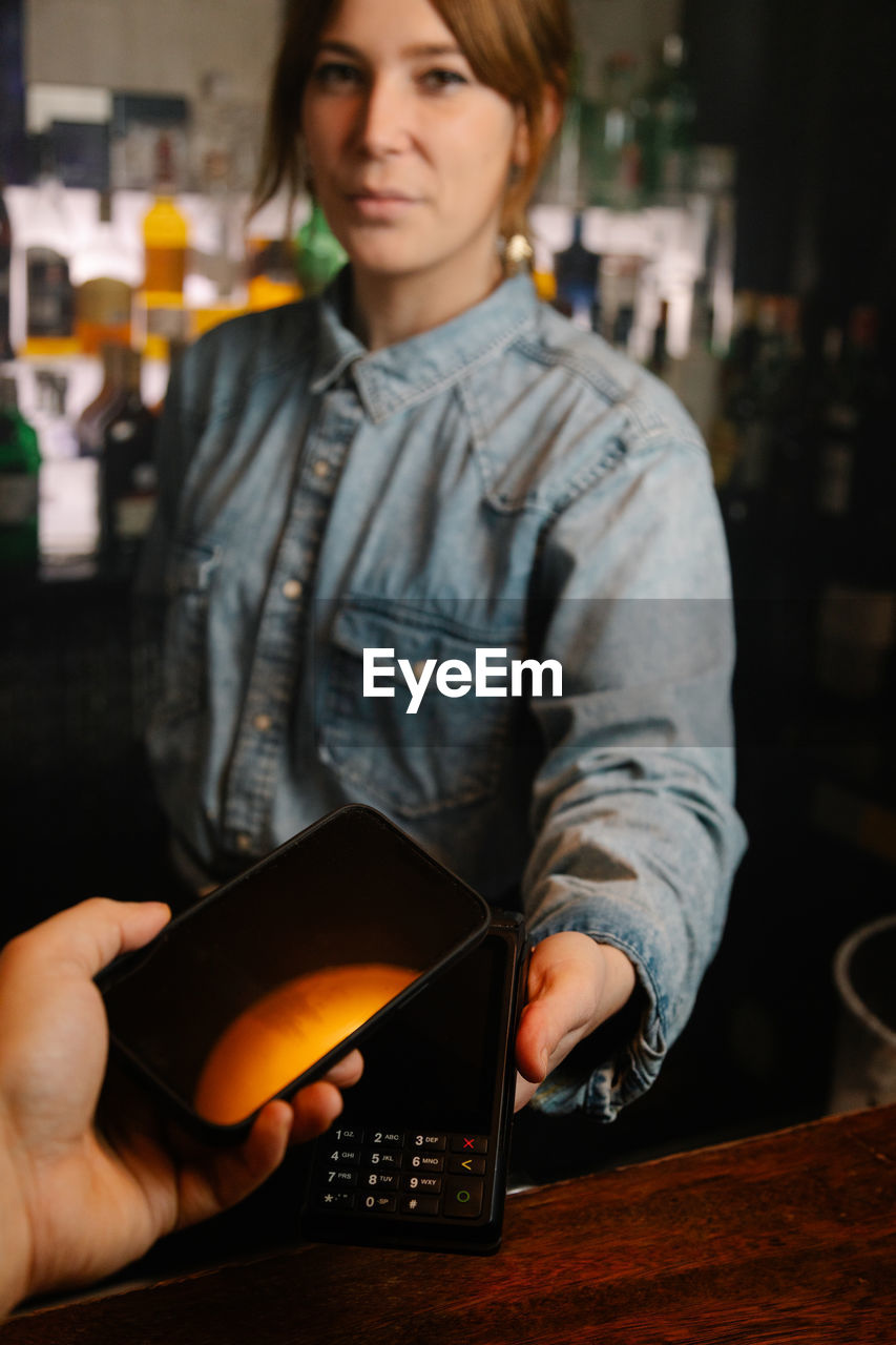 Crop anonymous client with making contactless payment by modern smartphone on terminal in bar near female bartender looking at camera