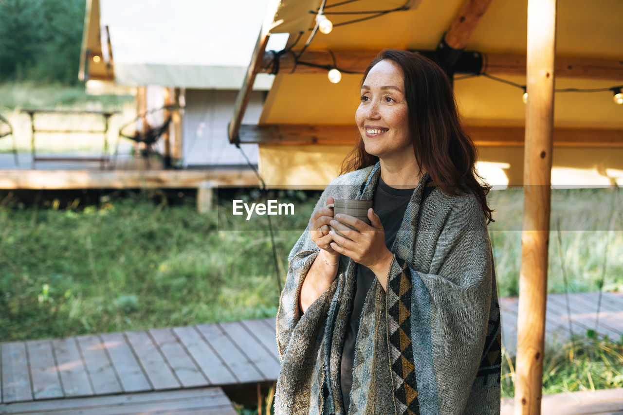 Young brunette woman in poncho drinking tea and relaxing in glamping in nature