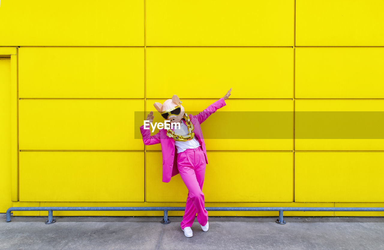 Woman wearing vibrant pink suit, pig mask and large golden chain posing with raised arms in front of yellow wall