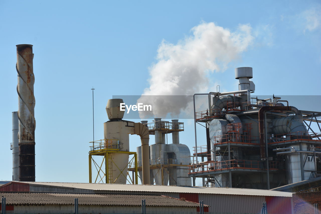 Low angle view of factory emitting smoke against blue sky