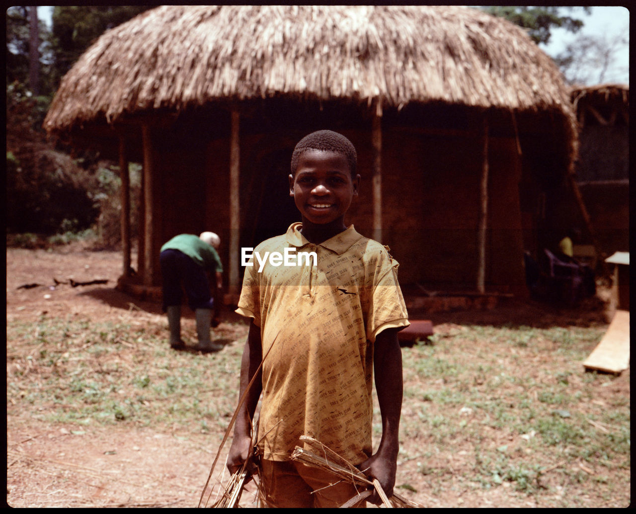 one person, hut, portrait, indigenous culture, architecture, village, men, thatched roof, adult, poverty, smiling, nature, person, looking at camera, standing, temple, building, traditional clothing, rural scene, front view, tribe, outdoors, transfer print, clothing, day, landscape, land, happiness, emotion