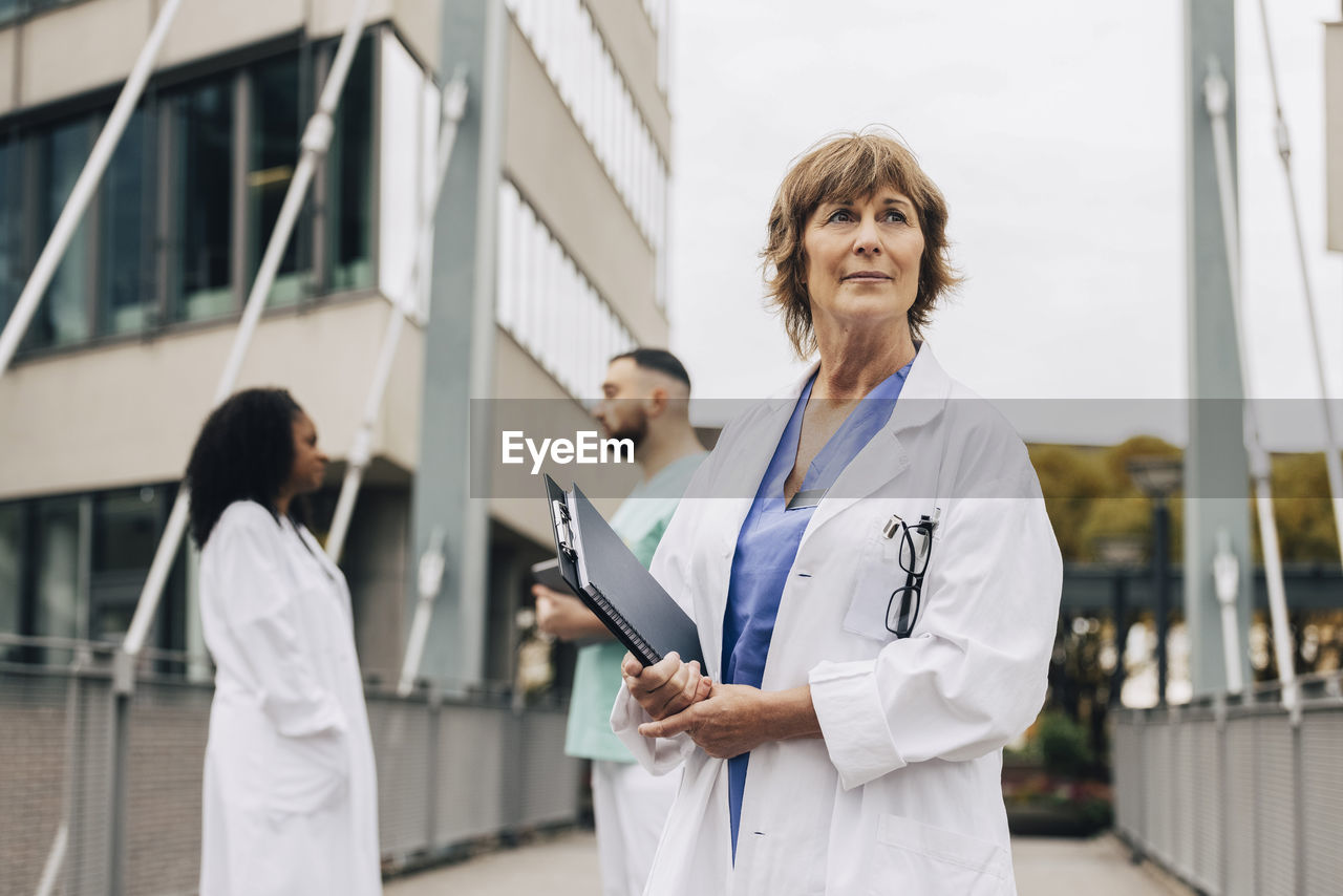 Mature female doctor looking away while holding clipboard and book outside hospital