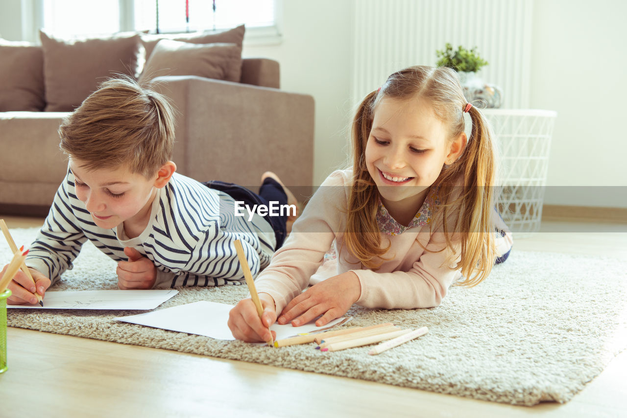 Siblings writing on paper while lying at home