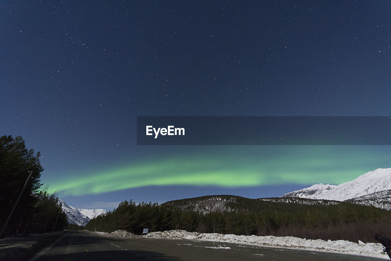 Road by snowcapped mountains against aurora borealis at night