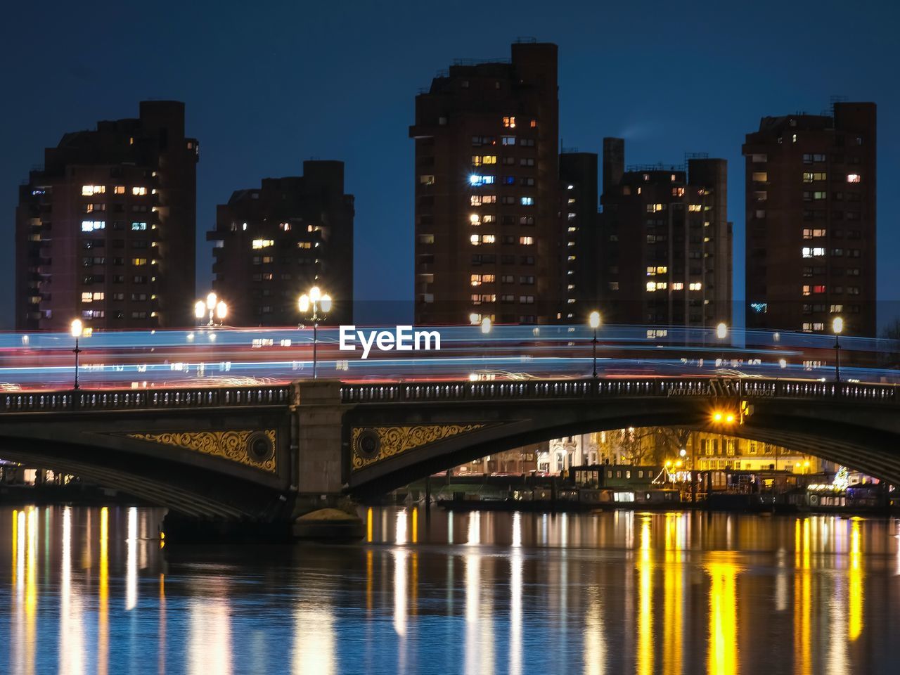 Illuminated bridge over river by buildings against sky at night