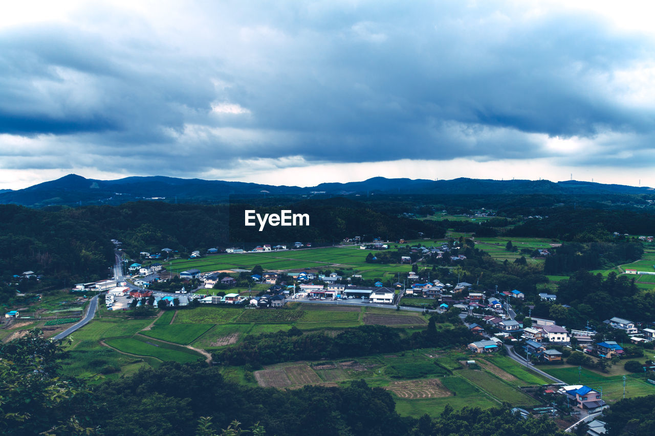 Scenic view of field and houses by mountains against cloudy sky