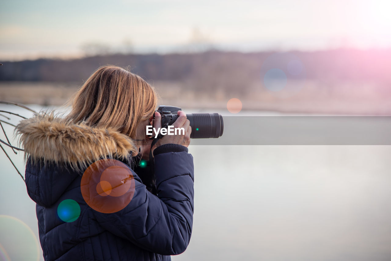 Side view of woman photographing with digital camera by lake during sunset