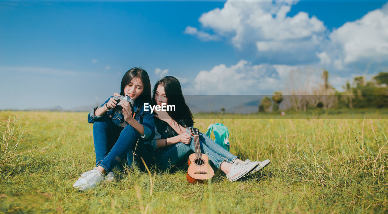 Young women sitting on grass while holding ukulele and camera against sky