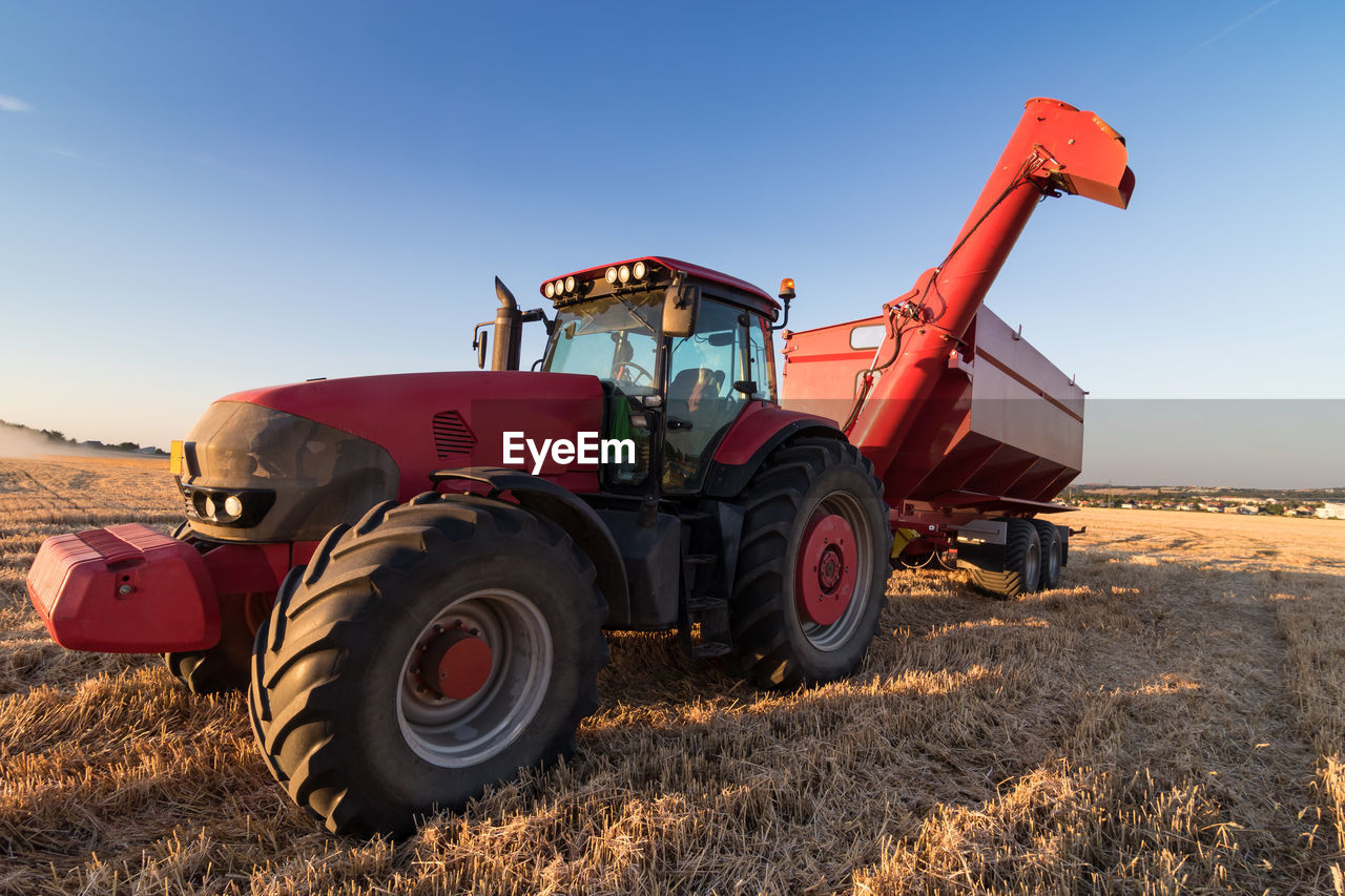 Agriculture tractor and tow trailer on a stubble field against clear sky