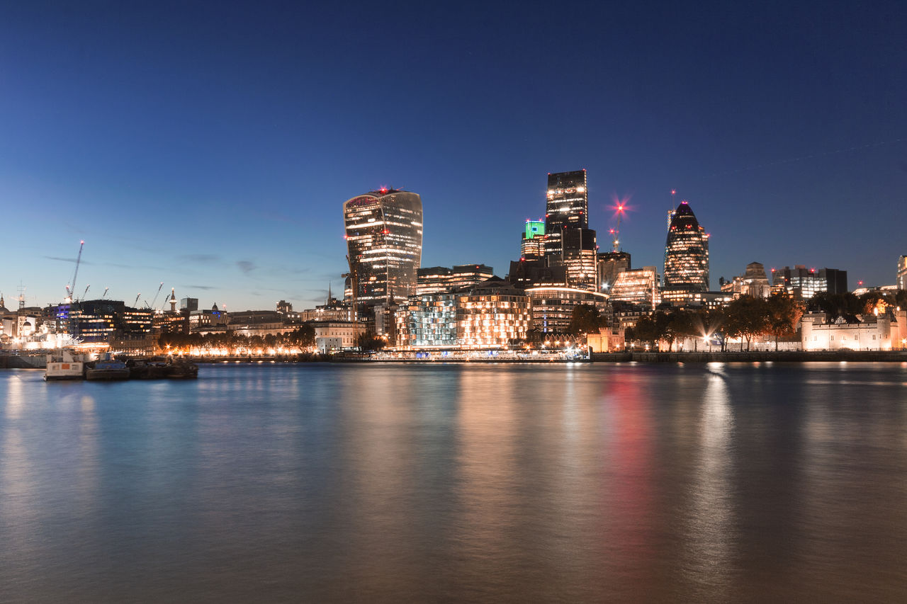 ILLUMINATED BUILDINGS BY RIVER AGAINST BLUE SKY