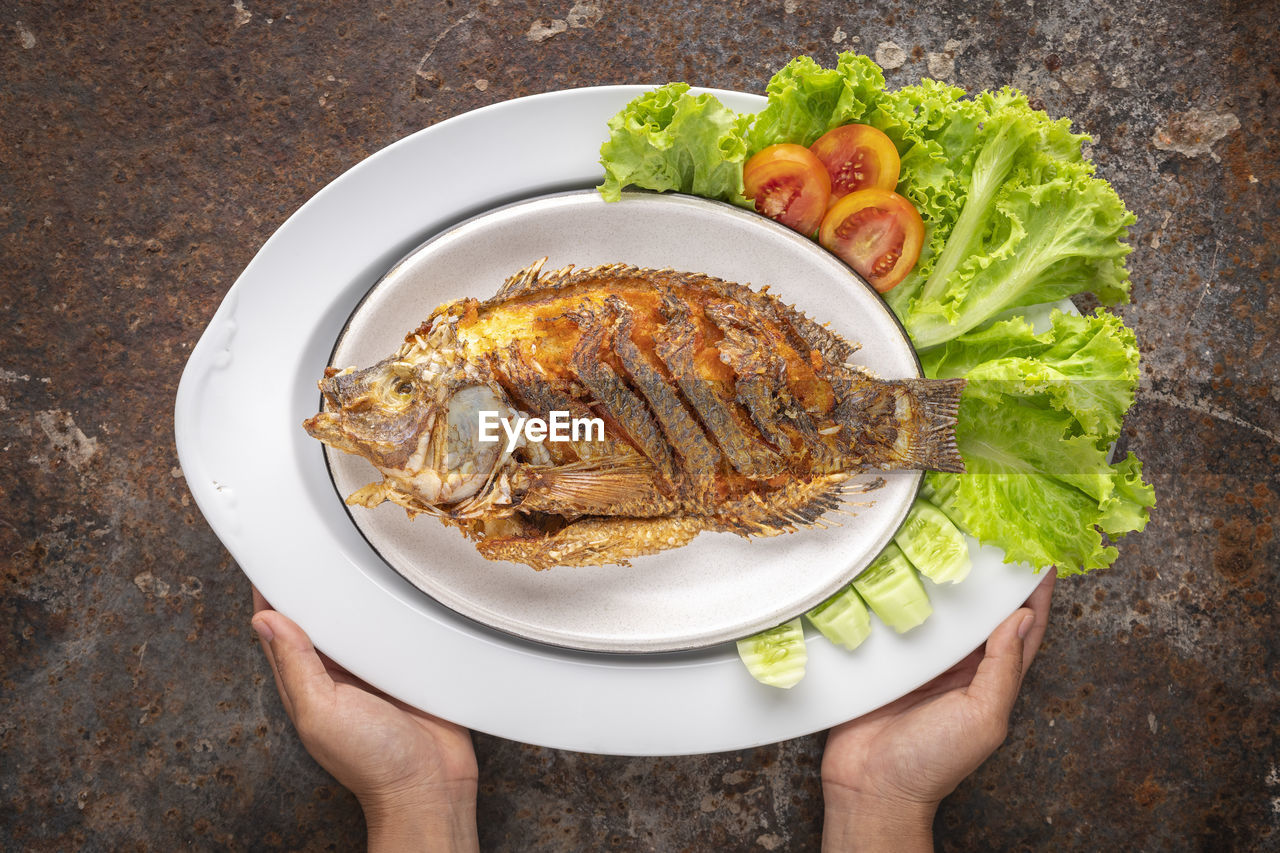 Fried nile tilapia fish with lettuce, tomato and cucumber in large oval ceramic plate served by hand