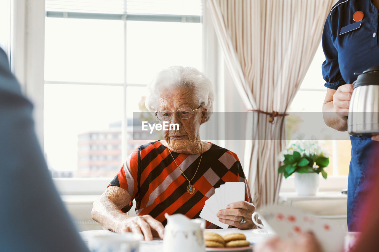 Senior woman playing cards with family while caretaker holding coffee pot at home