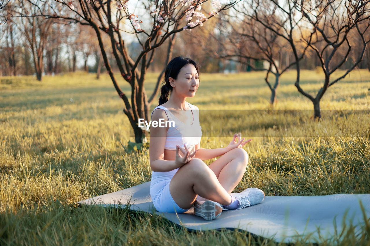 Asian woman practicing yoga sitting on grass, meditating with closed eyes under the tree.