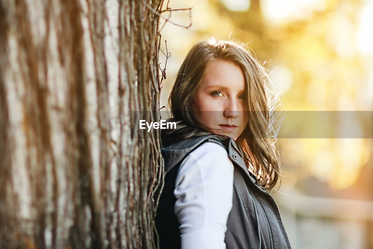 Portrait of beautiful young woman standing against tree trunk