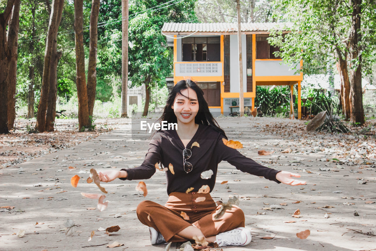 Portrait of smiling young woman throwing leaves while sitting on road in forest