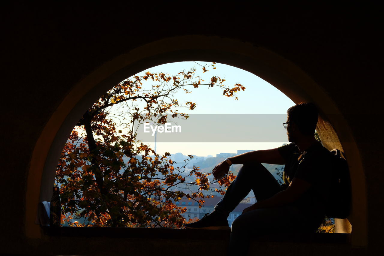 Silhouette man sitting by window against sky