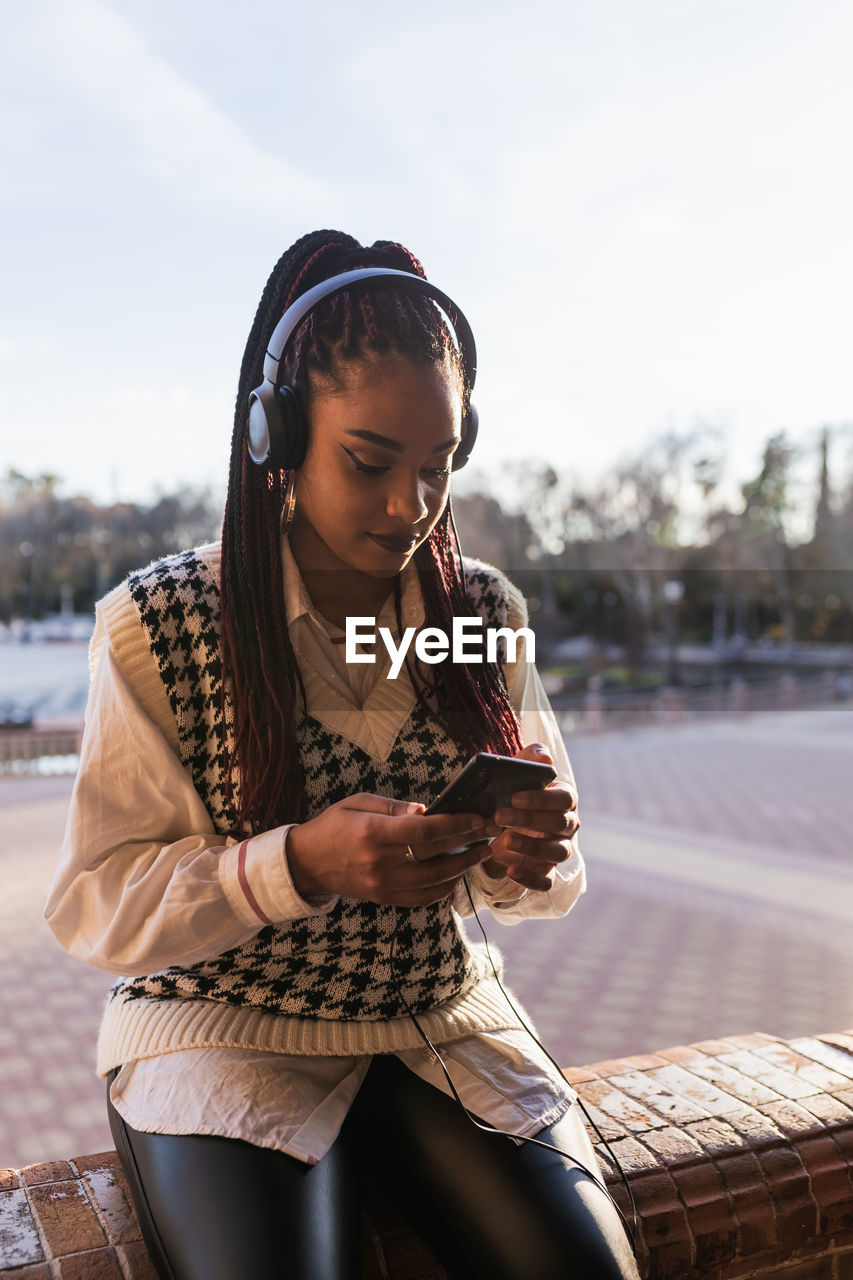 Charming african american female with braids sitting on stone fence on plaza de espana and enjoying music in headphones while browsing smartphone