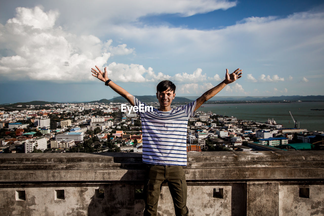 Portrait of smiling man with arms raised standing against cityscape by sea