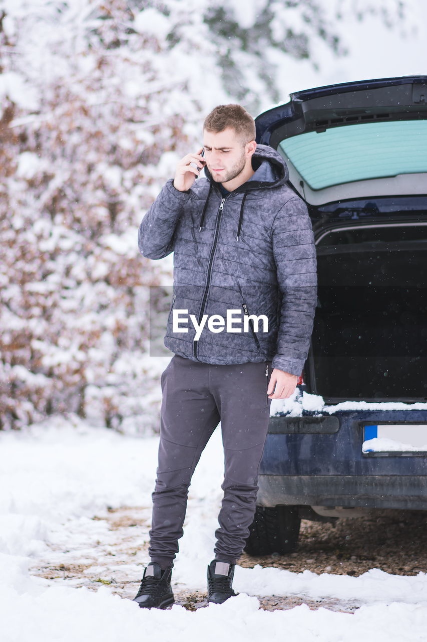 Young man using phone standing by broken down car in snow