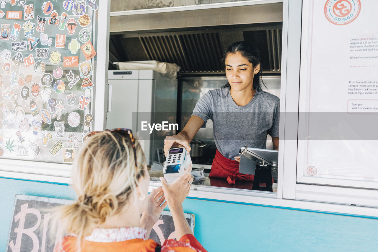 Young female owner standing giving credit card reader to blond customer at concession stand