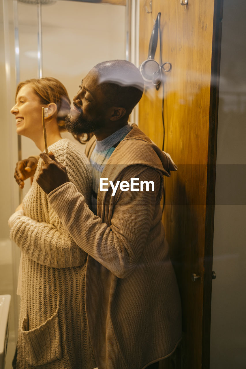 Side view of happy multiracial couple brushing teeth in bathroom at home