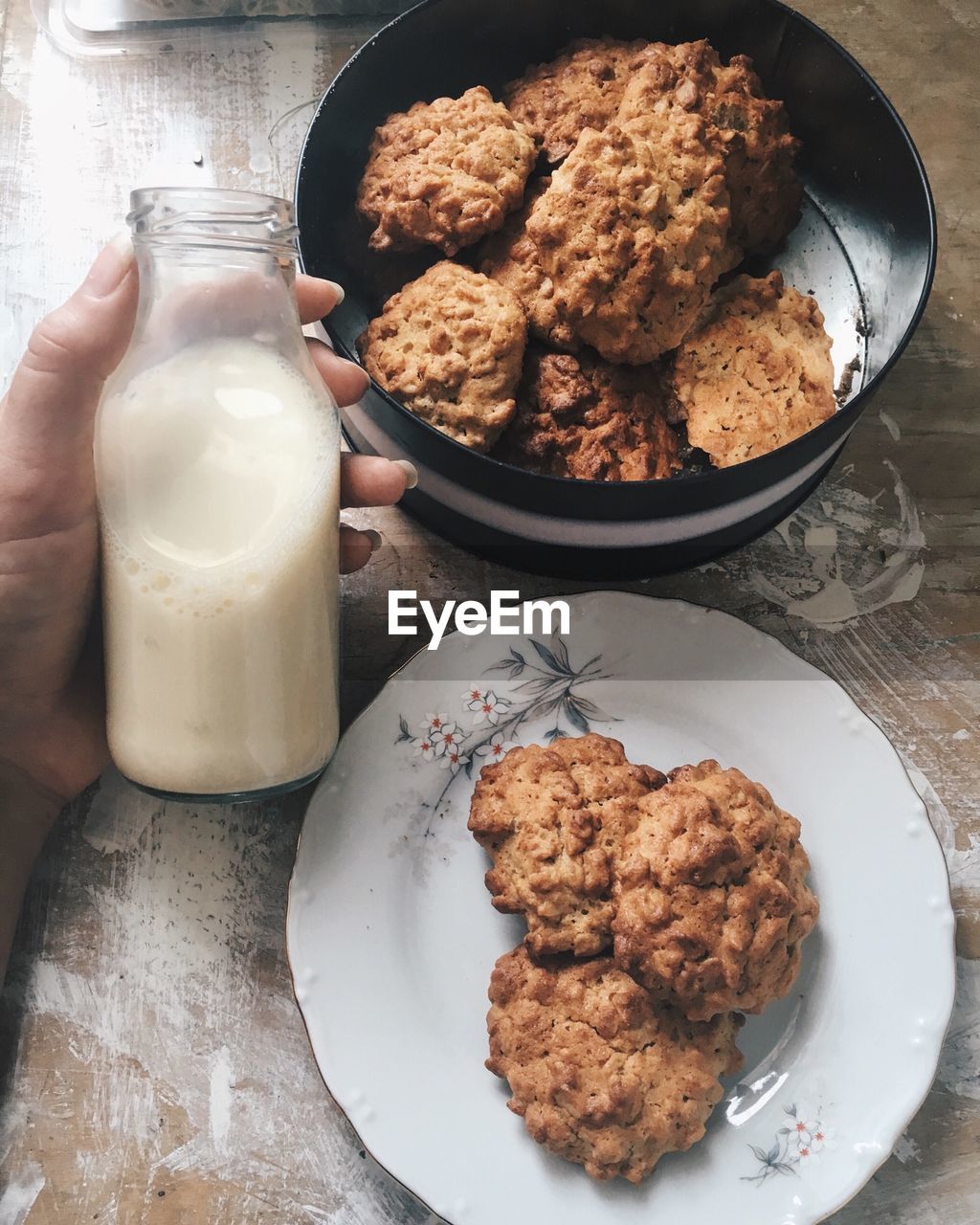 Cropped image of hand holding milk bottle by oatmeal cookies on table