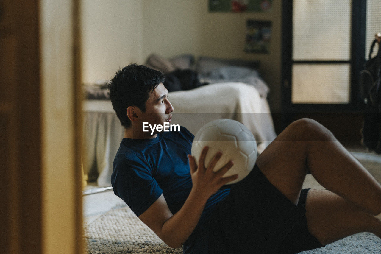 Ambitious teenage boy with soccer ball sitting in bedroom at home