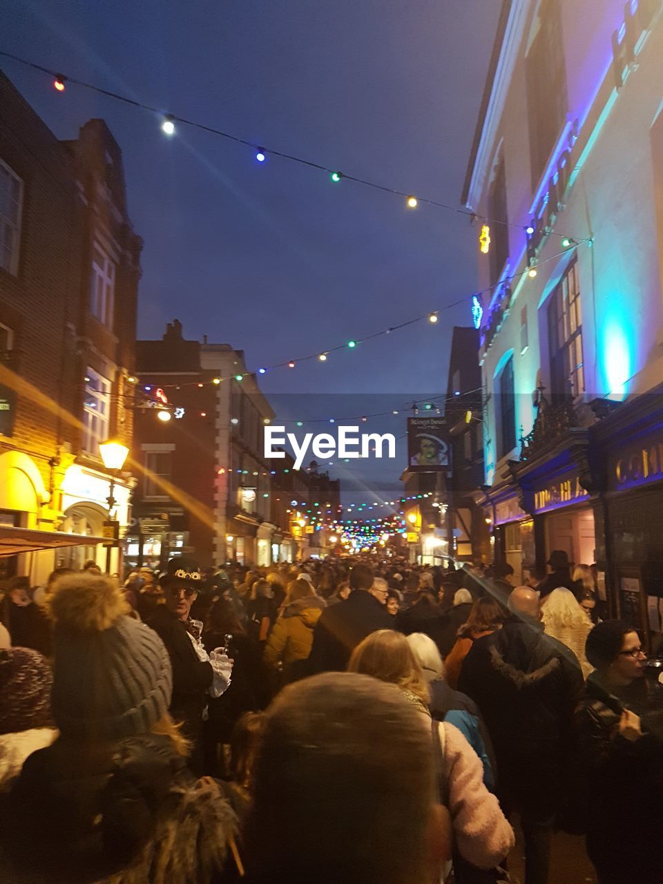 LOW ANGLE VIEW OF CROWD AT ILLUMINATED CITY AGAINST SKY