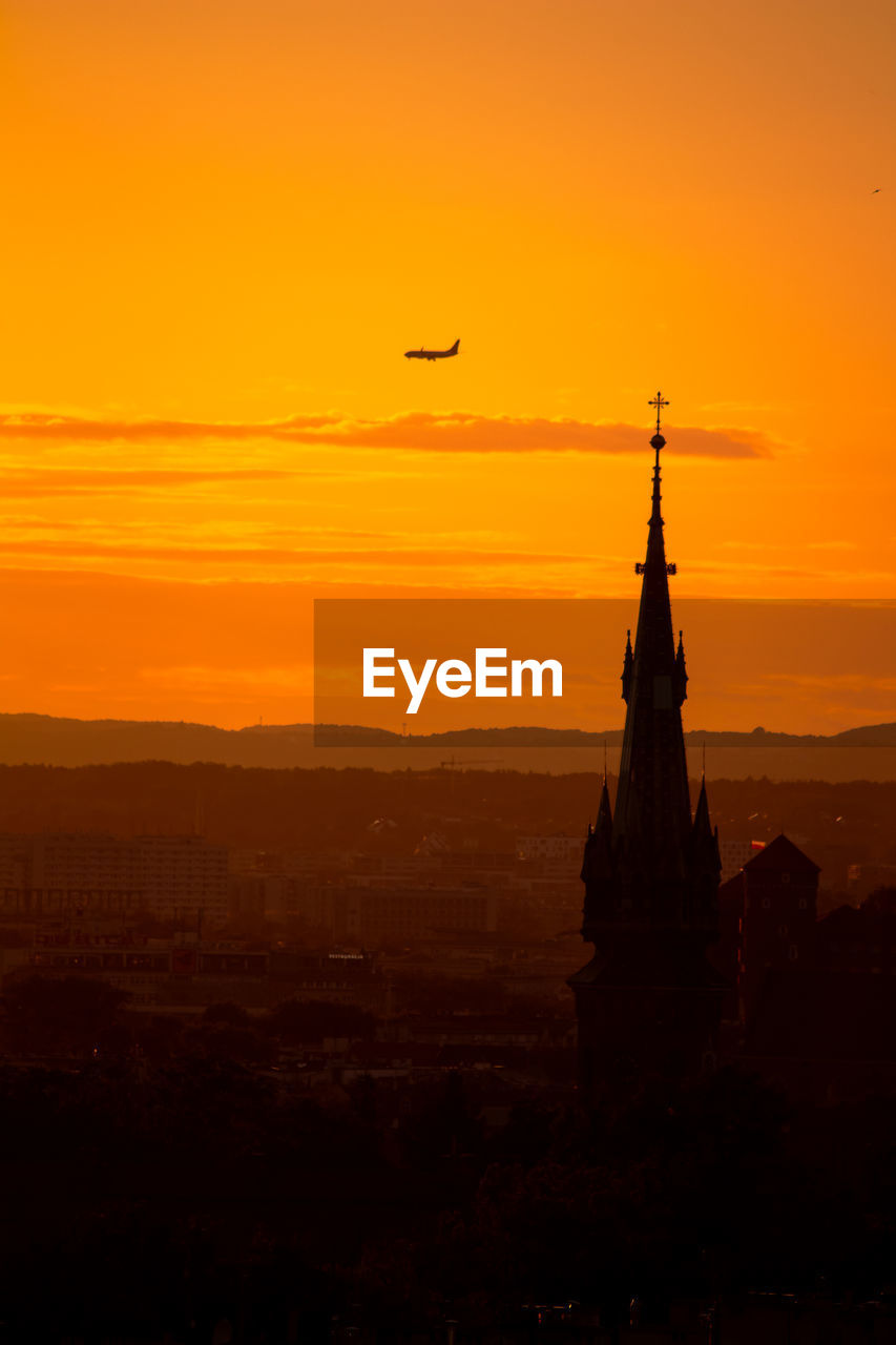 Orange sunset as a backdrop with a church tower and a plane passing through 