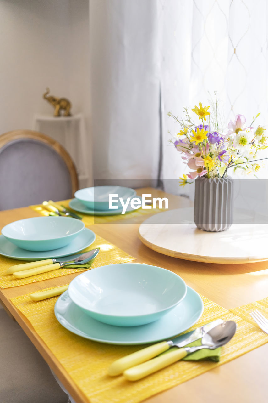 Dining set and flowers in vase on table, in brightly lit dining room.