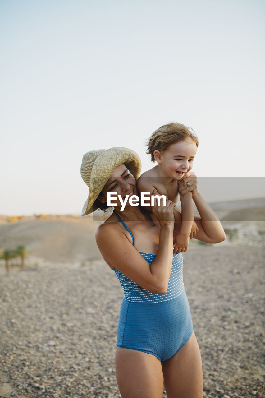 A mother wearing a hat holding her son up her shoulder in the desert