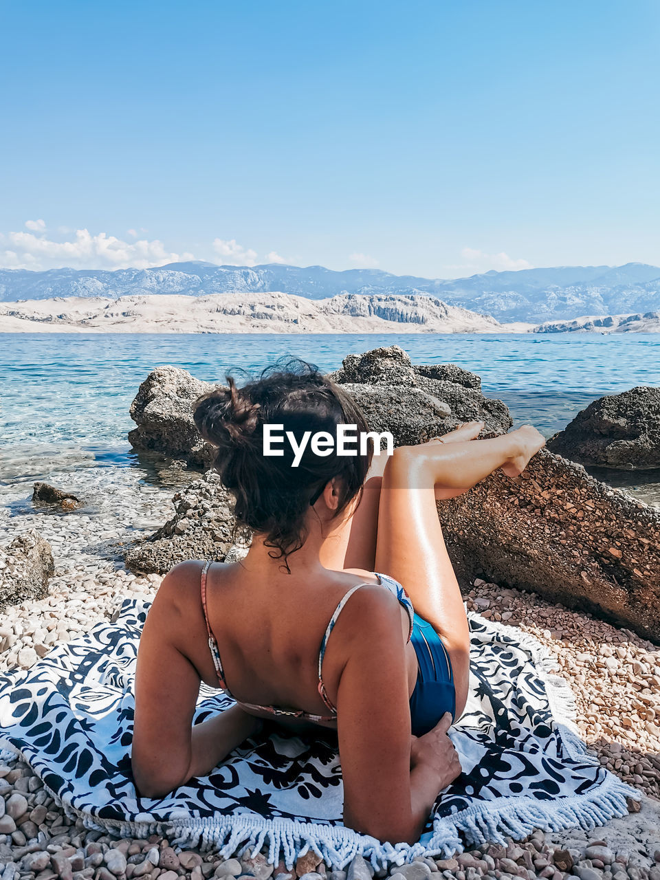 Rear view of beautiful young woman in bikini sunbathing on rocky beach.