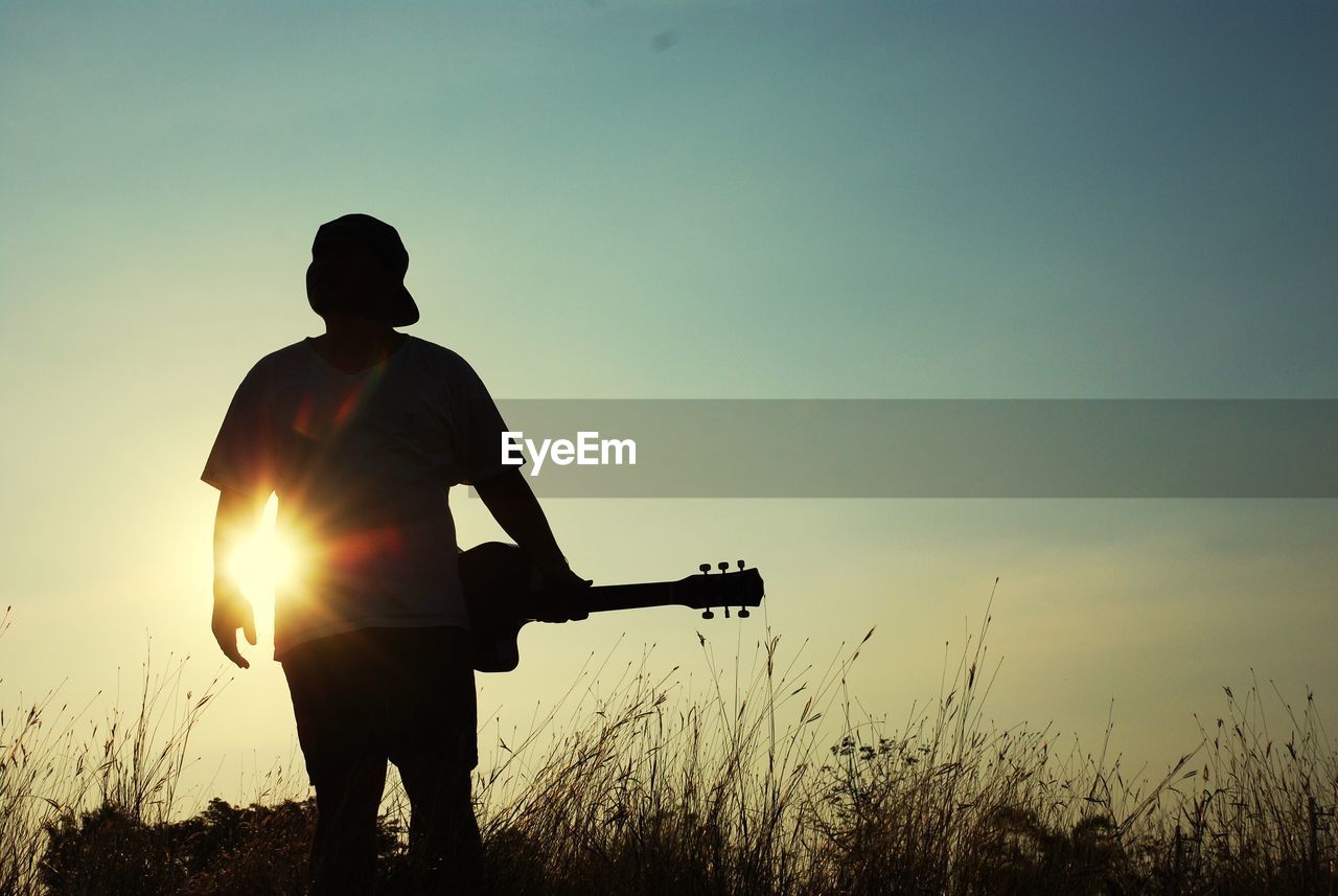 Silhouette man with guitar standing on field against sky during sunset