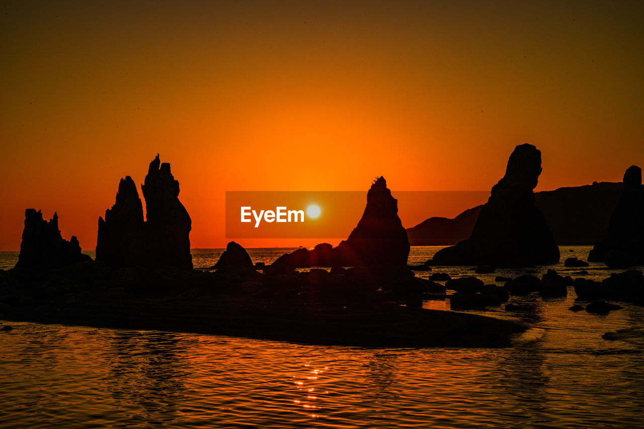 Silhouette rocks by sea against orange sky