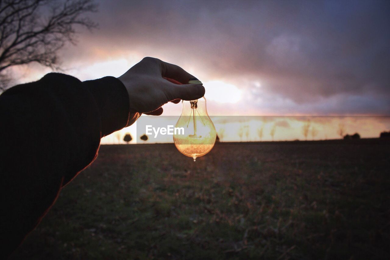 Close-up of hand holding light bulb against sky during sunset