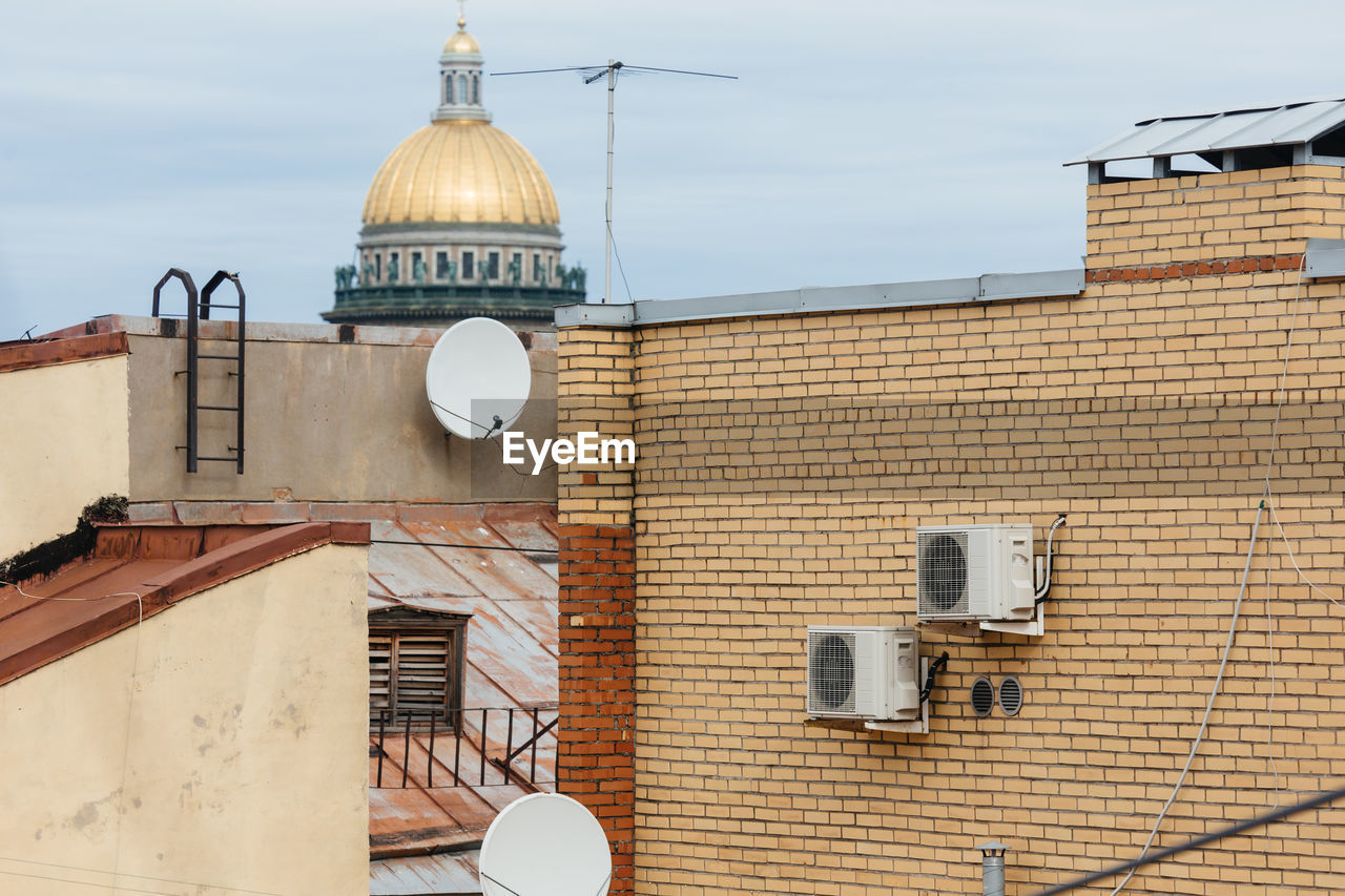 Two white air conditioners mounted on yellow brick wall and two sattelite dishes for television. 