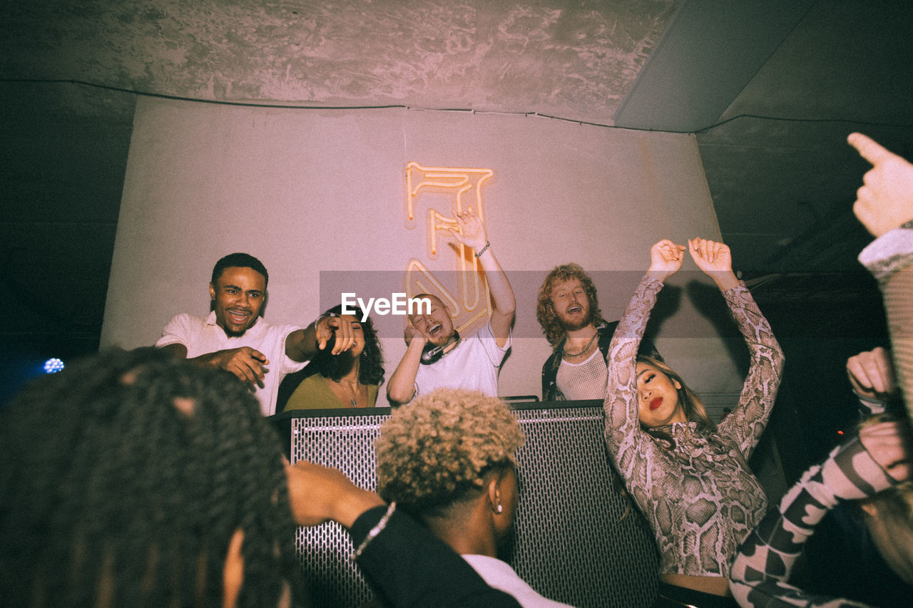 Happy young multiracial men and women dancing around dj at illuminated nightclub