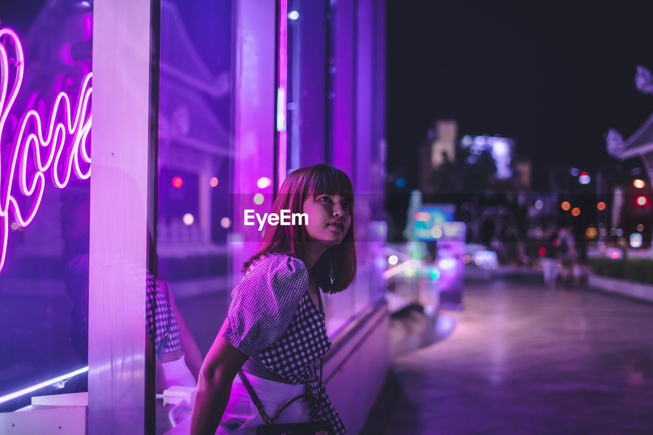 Young woman standing by illuminated building in city at night