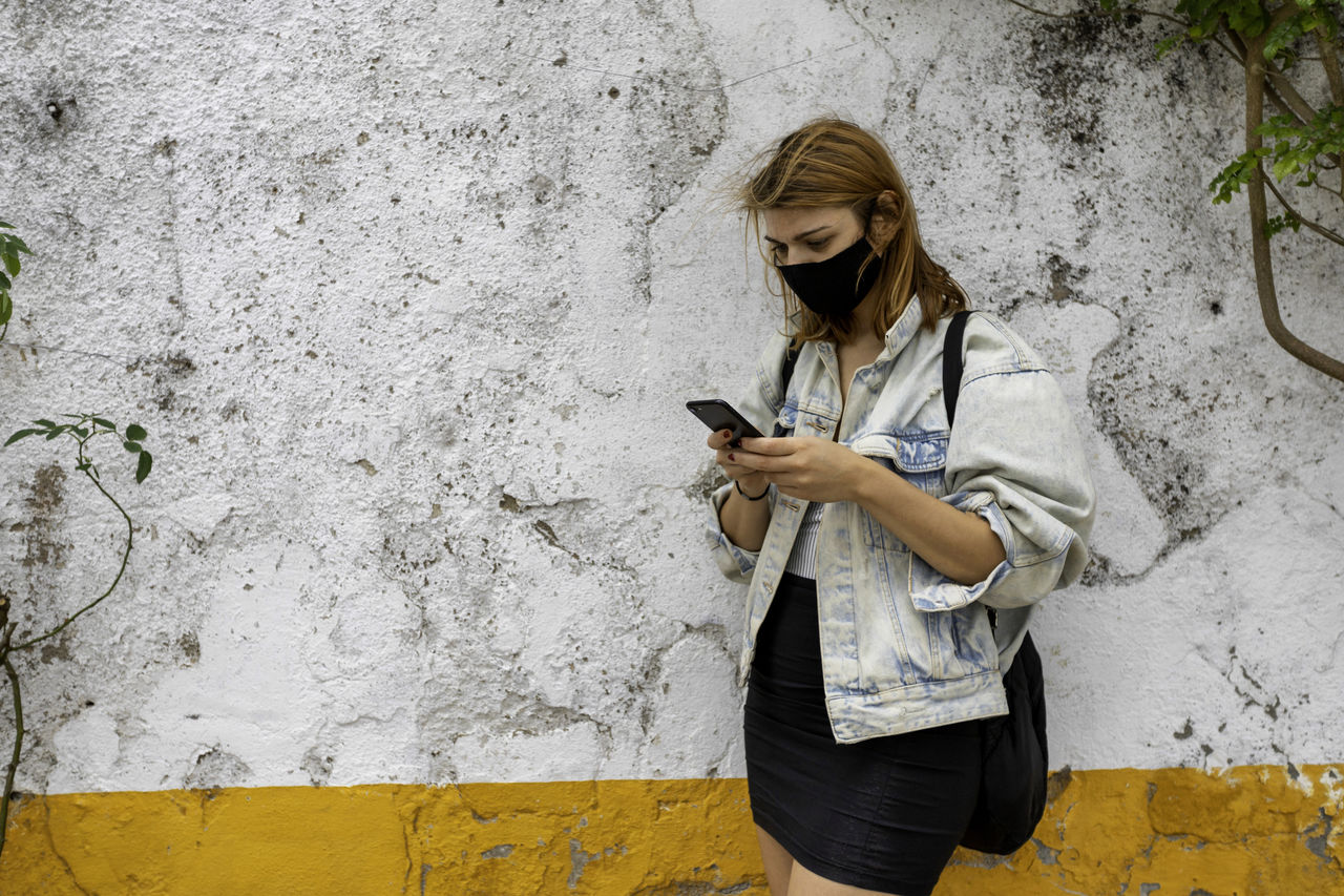 Smart woman with mask using mobile phone while standing by wall